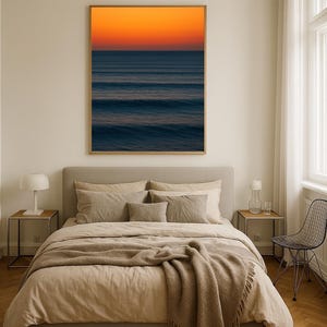 May include: A framed art print of an ocean scene with a vibrant orange and blue gradient. The artwork hangs above a bed with beige bedding and a brown throw blanket. The room has a neutral color palette with a modern aesthetic.