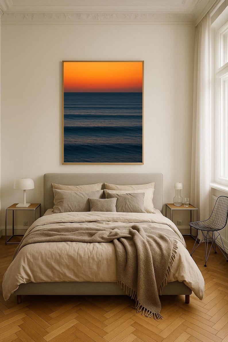 May include: A framed art print of an ocean scene with a vibrant orange and blue gradient. The artwork hangs above a bed with beige bedding and a brown throw blanket. The room has a neutral color palette with a modern aesthetic.