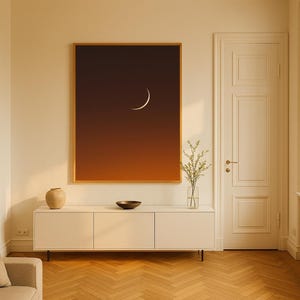 May include: A framed art print featuring a crescent moon against a gradient of orange and brown. The artwork hangs above a white cabinet with a vase, bowl, and plant. The room has a wooden floor and a beige sofa.