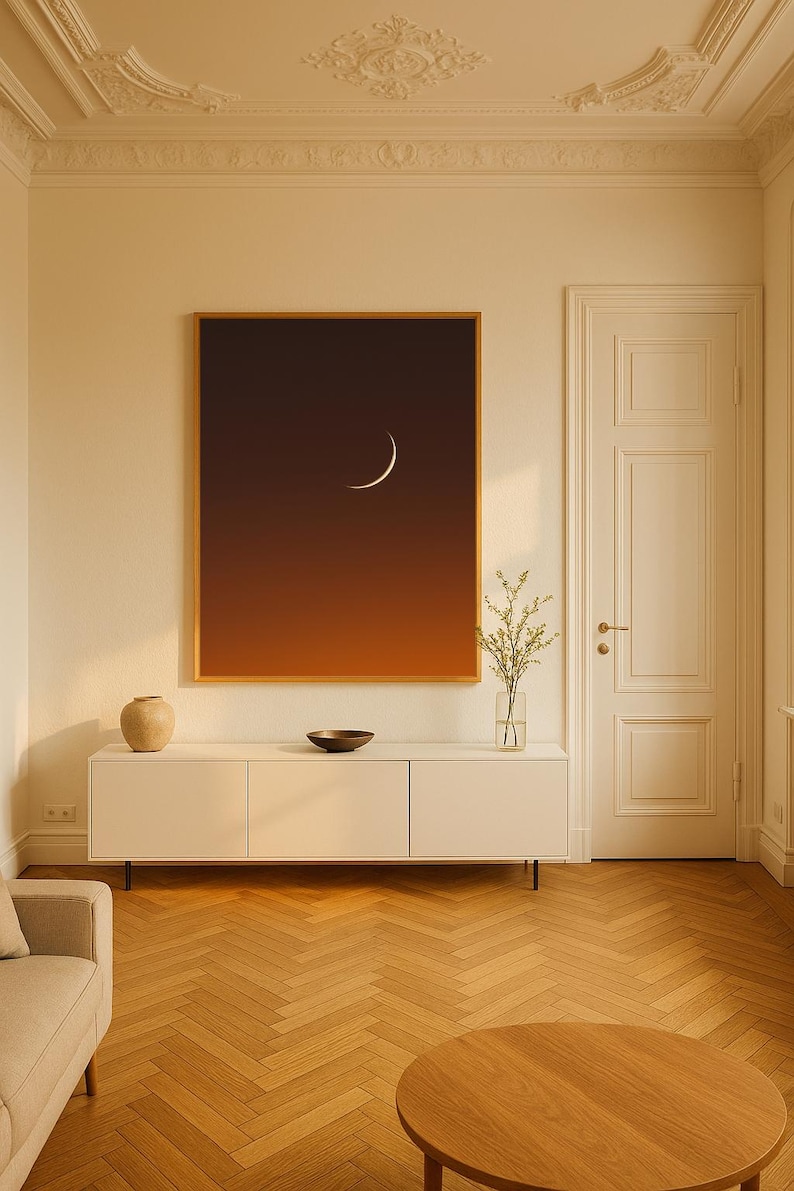 May include: A framed art print featuring a crescent moon against a gradient of orange and brown. The artwork hangs above a white cabinet with a vase, bowl, and plant. The room has a wooden floor and a beige sofa.