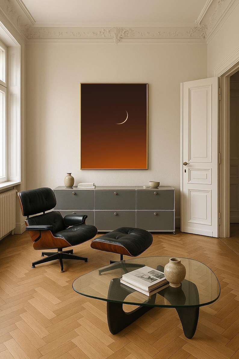 May include: Interior shot featuring a modern living room with a black leather chair and ottoman. A glass coffee table holds a book and vase. A framed art piece with a crescent moon hangs above a gray cabinet. The room has hardwood floors.