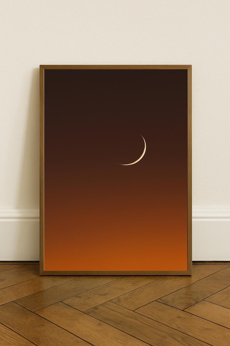 May include: A framed art print featuring a crescent moon against a gradient background of orange and brown hues. The artwork is displayed in a wooden frame, creating a minimalist aesthetic. The print is a celestial-themed home decor item.