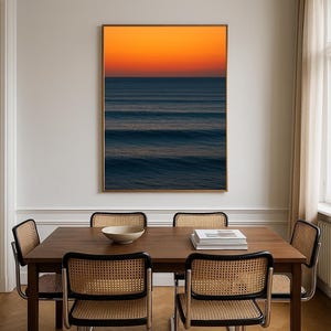 May include: A dining room scene featuring a dark wood table and four chairs with woven cane backs. A framed artwork of an ocean sunset hangs on the wall. A white bowl and books are on the table. The room has a neutral color palette.
