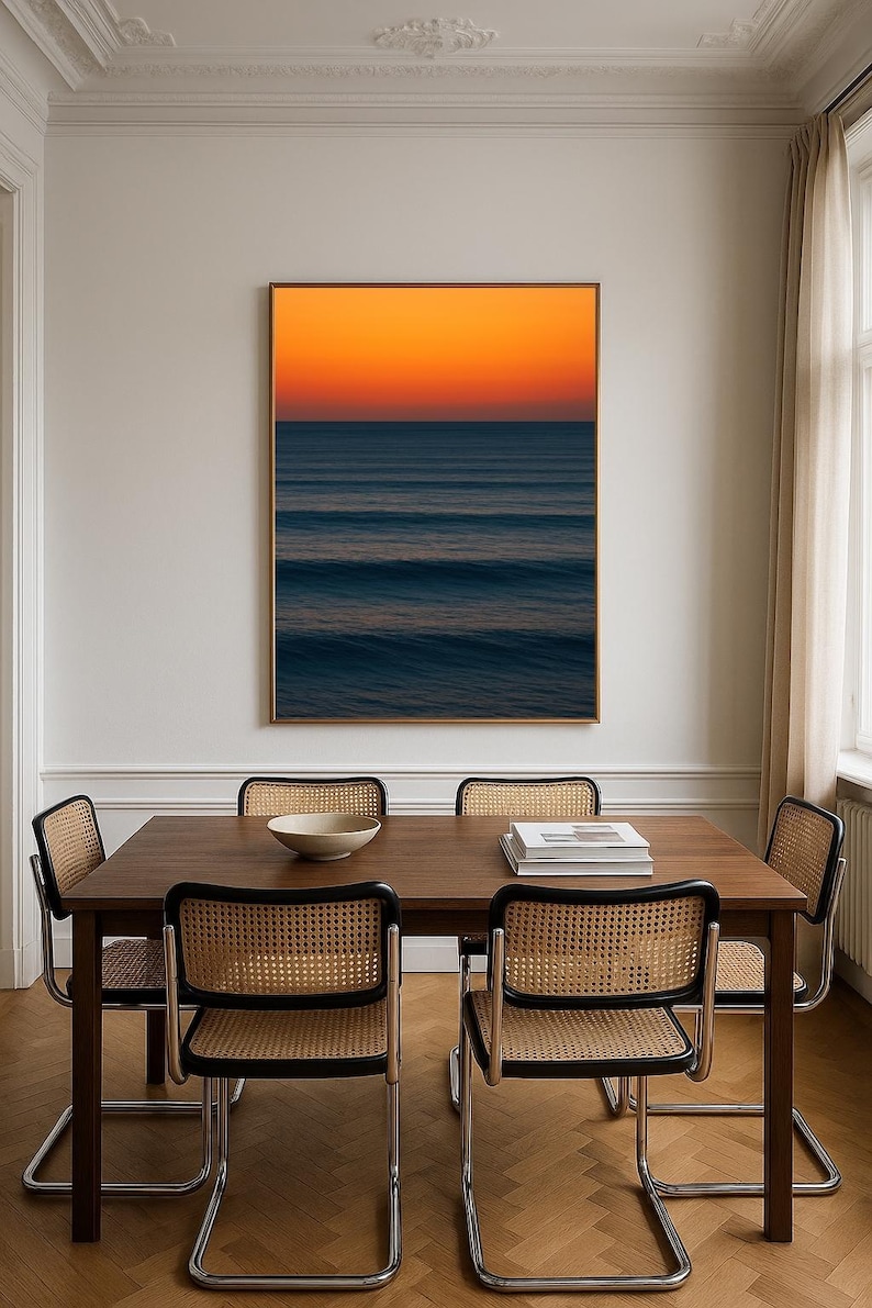 May include: A dining room scene featuring a dark wood table and four chairs with woven cane backs. A framed artwork of an ocean sunset hangs on the wall. A white bowl and books are on the table. The room has a neutral color palette.