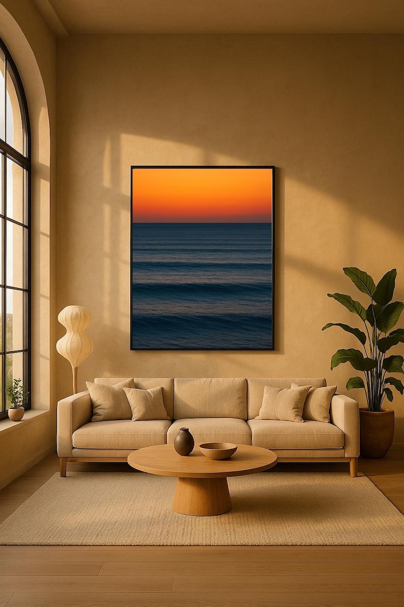 May include: A living room with a beige sofa, round wooden coffee table, and a large framed artwork depicting an ocean sunset. A floor lamp and potted plant add to the decor. The room has a neutral color palette.