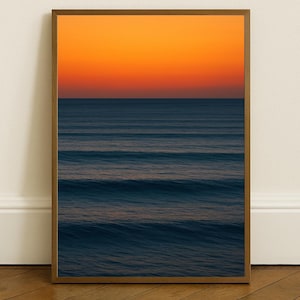 May include: A framed print featuring a seascape with a gradient sky of orange and yellow hues above a dark blue ocean. The water displays gentle waves. The wooden frame complements the warm tones of the artwork, creating a serene atmosphere.