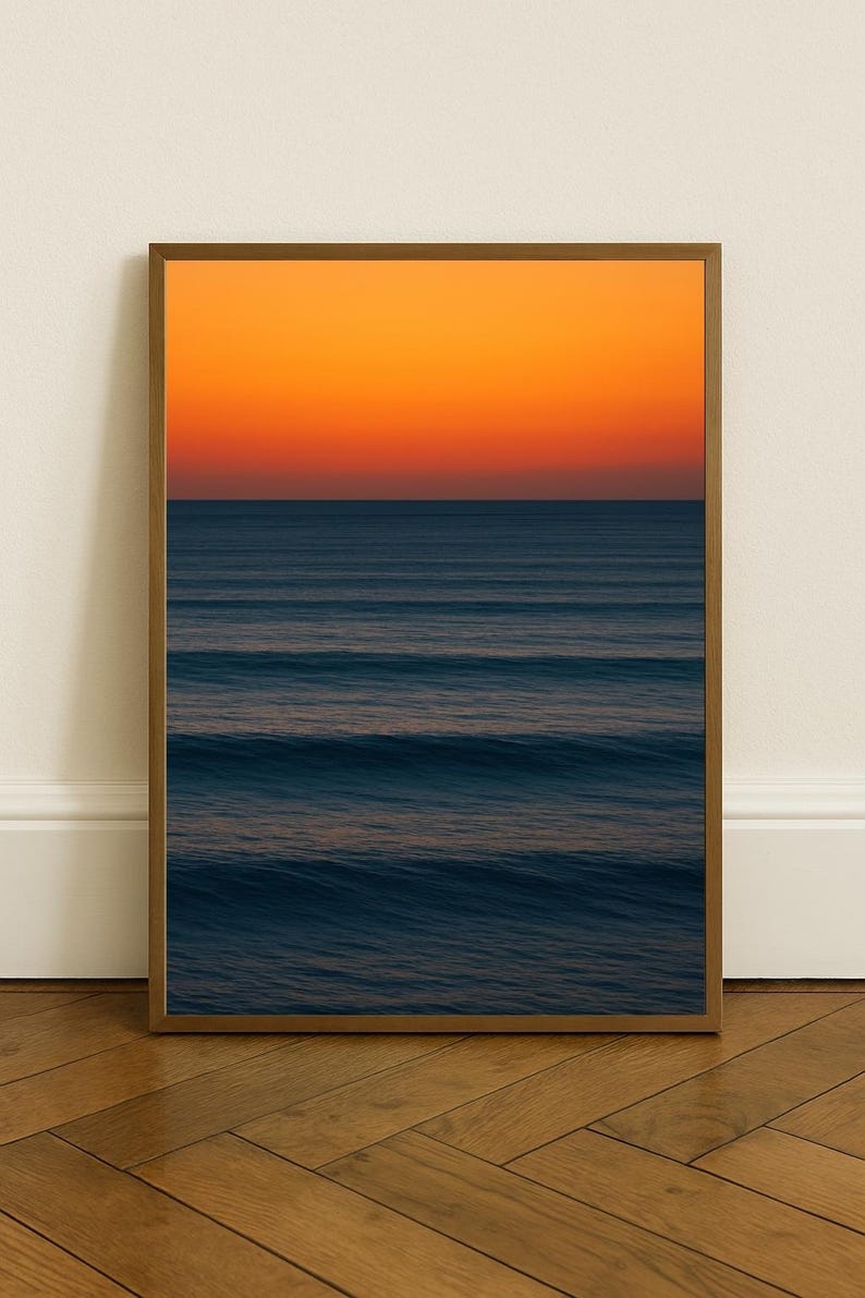May include: A framed print featuring a seascape with a gradient sky of orange and yellow hues above a dark blue ocean. The water displays gentle waves. The wooden frame complements the warm tones of the artwork, creating a serene atmosphere.