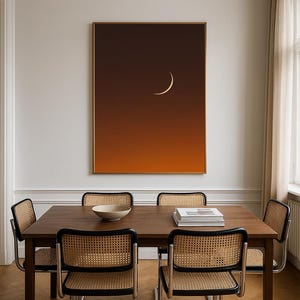 May include: A dining room scene featuring a dark wood table and four chairs with woven cane backs. A large framed artwork depicts a crescent moon against a gradient orange and brown background. A white bowl and books are on the table.