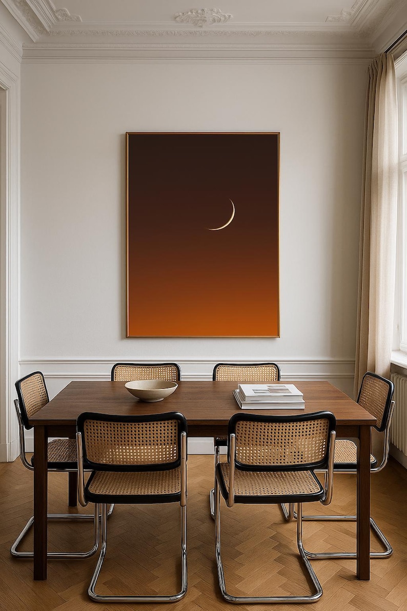 May include: A dining room scene featuring a dark wood table and four chairs with woven cane backs. A large framed artwork depicts a crescent moon against a gradient orange and brown background. A white bowl and books are on the table.