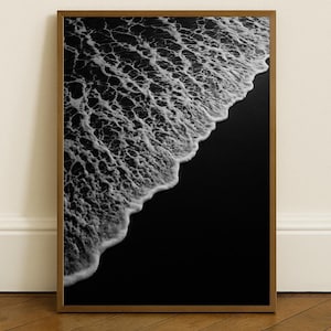 May include: A framed black and white photograph of a beach scene. The image captures the foamy white crest of a wave meeting the dark sand. The wooden frame adds a natural touch to the minimalist coastal artwork.