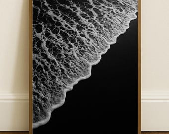 May include: A framed black and white photograph of a beach scene. The image captures the foamy white crest of a wave meeting the dark sand. The wooden frame adds a natural touch to the minimalist coastal artwork.