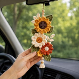 Könnte beinhalten: Ein hängendes Auto-Accessoire mit einer Ansammlung von gehäkelten Blumen in Gelb-, Creme- und Rostorangetönen, akzentuiert durch grüne gehäkelte Blätter. Das Arrangement hängt an einer Schnur mit Holzperlen und einer kleinen cremefarbenen Quaste.