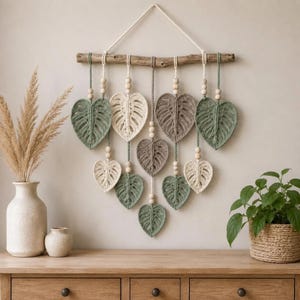 Op de afbeelding: Een wandhanger met een horizontale houten tak waaraan meerdere macramé monstera-bladeren hangen in tinten saliegroen, crème en taupe. Elk blad hangt aan koorden die zijn versierd met kleine houten kralen in een gelaagde compositie.