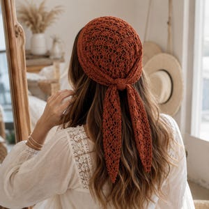 May include: A rust-colored crochet headscarf featuring an intricate openwork fan pattern. The headscarf is tied at the back of the head with two long, pointed triangular tails hanging down, crafted from a lightweight, textured yarn material.