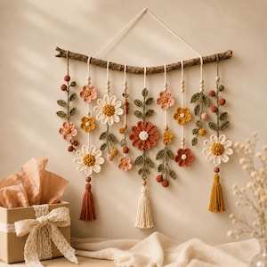 May include: A wall hanging featuring a natural wooden branch suspending multiple crocheted floral and leaf strands. The arrangement includes white, yellow, pink, and rust-colored flowers, green foliage, wooden beads, and three hanging yarn tassels.
