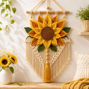 May include: A macrame wall hanging featuring a large sunflower design with a dark brown center and layered yellow and orange petals. The piece includes green leaf accents, cream-colored knotted cord patterns, wooden beads, and a long yellow tassel.