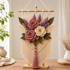 May include: A macrame wall hanging featuring a wooden dowel, cream-colored knotted cord, and a central arrangement of fabric flowers in shades of dusty rose and cream, accented with purple sprigs, green leaves, and a dusty rose tassel.