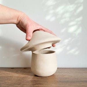 May include: A beige ceramic mushroom-shaped container with a lid being lifted. The container has a rounded base and a wide, flat lid. The background is a plain white wall and a wooden surface.