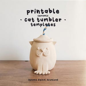 May include: A beige ceramic cat-shaped tumbler with a hat and a blue and white striped straw. The tumbler has a cat face design with two dots for eyes and a dotted nose. Text on the image reads "printable ceramic cat tumbler templates" and "instant digital download."