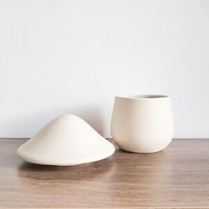 May include: Two off-white ceramic pieces sit on a wooden surface. One is a rounded, cone-shaped object, and the other is a small, rounded cup. The objects are simple and minimalist, with a matte finish.