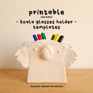 May include: A ceramic koala glasses holder with the text "printable ceramic - koala glasses holder - templates". The holder is white and has a slot for glasses and several colored markers.