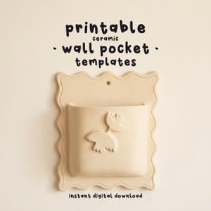 Puede incluir: Un bolsillo de pared de cerámica beige con borde festoneado y un pequeño orificio para colgar. El frente presenta un diseño en relieve de dos pájaros estilizados. Sobre el objeto, el texto dice printable ceramic wall pocket templates.