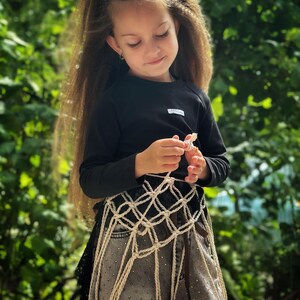 May include: A black long-sleeved shirt with a small white rectangular label on the chest, paired with grey denim pants featuring a subtle metallic stud pattern and an overlay of beige macrame netting with a knotted diamond-shaped design.
