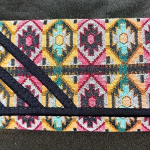 May include: A rectangular wallet with a colorful geometric pattern in pink, yellow, teal, and black. The wallet has a black wrist strap and black diagonal accents on the front.