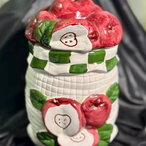 May include: A white ceramic cookie jar with a basket weave design, adorned with red apple accents and green leaves. The lid is topped with red apples and a green and white checkered band.