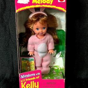 Puede incluir: Una muñeca vintage "Bunny Melody" en su embalaje original. La muñeca lleva un disfraz de conejito rosa con una barriga blanca y orejas de conejo. La caja presenta el texto "Adventures with Li'l Friends of Kelly" y una advertencia sobre piezas pequeñas.