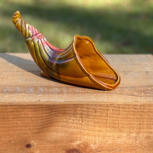 May include: A ceramic cornucopia vase in shades of brown, yellow, and red. The horn-shaped vase is resting on a wooden surface. The vase has a glossy finish and is likely vintage.