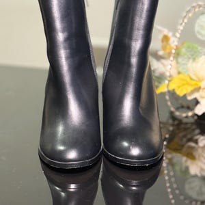 Könnte beinhalten: Ein Paar schwarze Leder-Ankle Boots mit Blockabsatz. Die Stiefel haben einen seitlichen Reißverschluss und eine runde Spitze. Die Stiefel sind etwa 15 cm hoch.