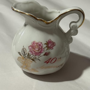 May include: A small, white ceramic pitcher with a gold rim and handle. The pitcher is decorated with pink roses and the text "40th Anniversary" in red. The handle has a decorative swirl design.