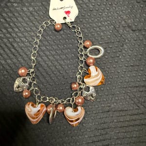 May include: A silver-tone charm bracelet featuring brown and white swirled heart-shaped beads, metallic heart-shaped padlock charms, and round brown beads. The bracelet is attached to a white display card with the text Fashion Jewelry printed on it.