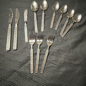 May include: A collection of eleven silver-toned metal utensils featuring a repeating geometric pattern on the handles. The set includes two dinner knives, one butter knife, five spoons of varying sizes, and three forks, all with matching designs.