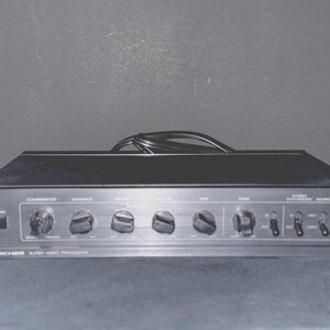 Op de afbeelding: Een zwarte Archer Super Video Processor met meerdere knoppen en schakelaars. Het voorpaneel heeft bedieningselementen met de labels "Comparator", "Enhance", "Stop" en "Fade". Aan de linkerkant zit een aan/uit-knop. Een zwarte kabel is aangesloten aan de bovenkant.