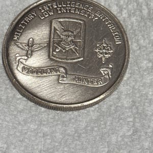 May include: A silver Military Intelligence Battalion coin. The coin features the text "MILITARY INTELLIGENCE BATTALION LOW INTENSITY" around the edge. The center has a crest with crossed swords and a banner that reads "VIGILANT HUNTER".
