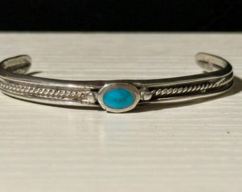 May include: A silver cuff bracelet featuring an oval turquoise stone. The bracelet has a twisted rope design along the edges. The turquoise stone is a vibrant blue colour. The bracelet is displayed on a light surface.