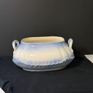 May include: A light blue and cream-colored ceramic serving bowl with two loop handles. The bowl has a scalloped rim and a rounded base. The interior is cream-colored, while the exterior fades from light blue to cream.