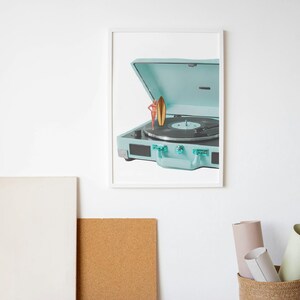 May include: A framed print featuring a vintage-style, light blue record player with its lid open. A small surfboard figurine stands on the record. The record player has a black record on the turntable. The image is set against a white background.