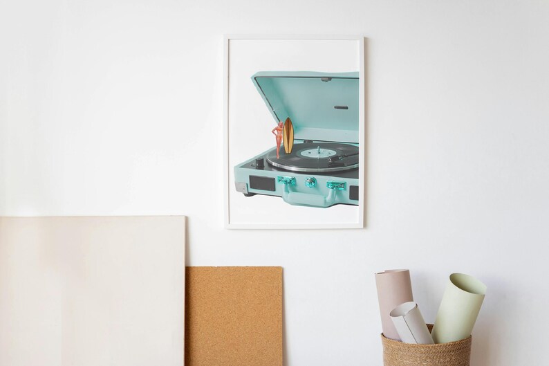 May include: A framed print featuring a vintage-style, light blue record player with its lid open. A small surfboard figurine stands on the record. The record player has a black record on the turntable. The image is set against a white background.