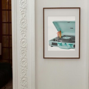 May include: A framed print featuring a vintage-style, teal record player with a vinyl record. The record player is open, and a small surfboard and figure are placed on top. The frame is brown, and the print is mounted on a white background.