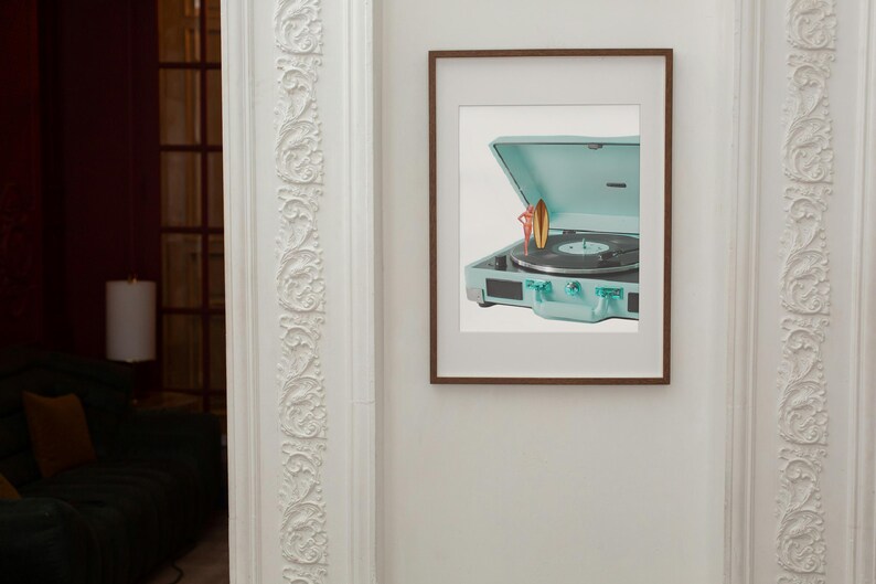 May include: A framed print featuring a vintage-style, teal record player with a vinyl record. The record player is open, and a small surfboard and figure are placed on top. The frame is brown, and the print is mounted on a white background.