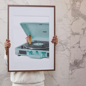 May include: A framed print featuring a vintage-style, light blue record player with a black vinyl record. A miniature figure of a person with a surfboard is standing on the record player. The frame is brown.