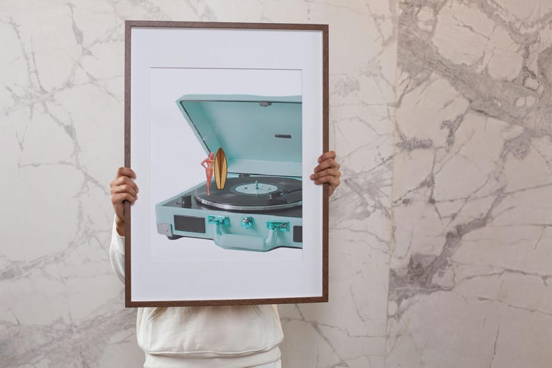 May include: A framed print featuring a vintage-style, light blue record player with a black vinyl record. A miniature figure of a person with a surfboard is standing on the record player. The frame is brown.