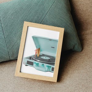 May include: A framed print featuring a vintage-style, light blue record player with a surfboard and a person. The record player has a black turntable and clear knobs. The frame is light brown and rests on a brown surface next to a teal pillow.
