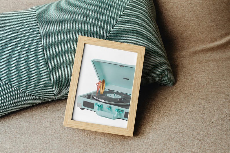 May include: A framed print featuring a vintage-style, light blue record player with a surfboard and a person. The record player has a black turntable and clear knobs. The frame is light brown and rests on a brown surface next to a teal pillow.