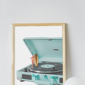May include: A framed print featuring a vintage-style, light blue record player with a miniature surfboard and a figurine of a person in a swimsuit. The record player is open, revealing a black vinyl record.