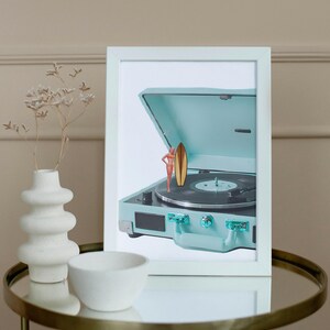 May include: A framed image featuring a vintage-style, light blue record player with a miniature surfer and surfboard. The record player is open, revealing the turntable. The image is set on a gold-rimmed table with a white vase and bowl.