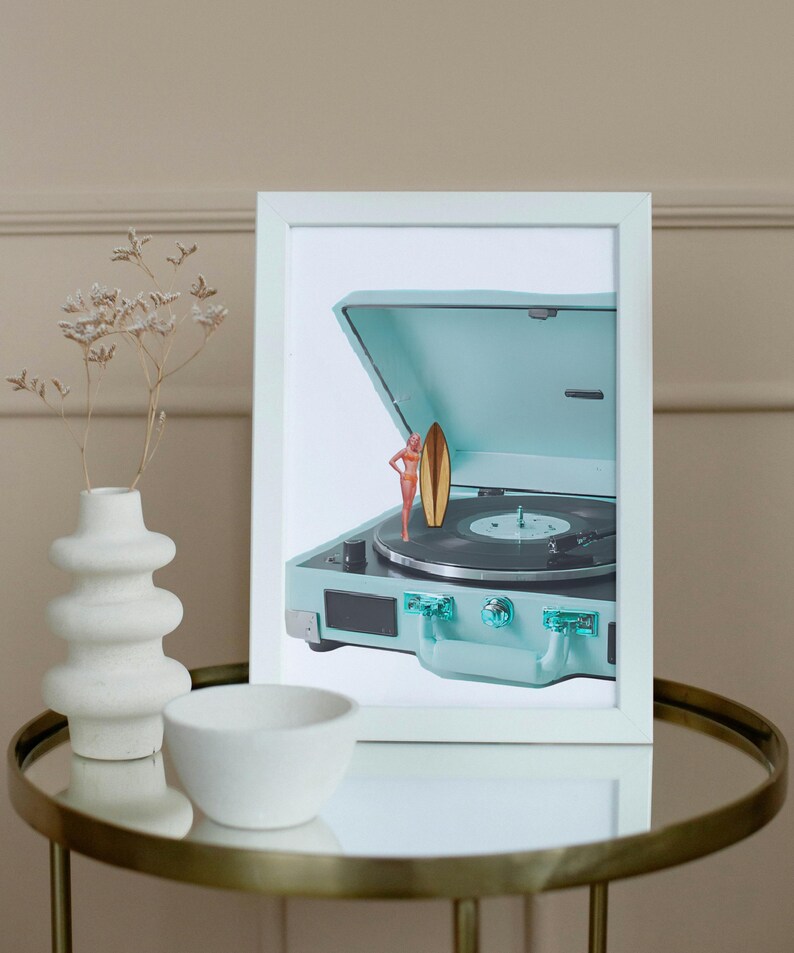 May include: A framed image featuring a vintage-style, light blue record player with a miniature surfer and surfboard. The record player is open, revealing the turntable. The image is set on a gold-rimmed table with a white vase and bowl.